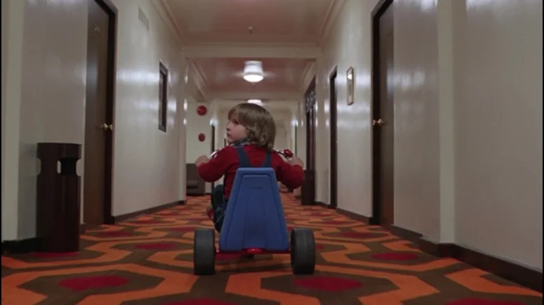 어린아이가 세발자전거를 타고 반복되는 Overlook Hotel 복도를 지나가는 낮은 시점의 영화 스틸